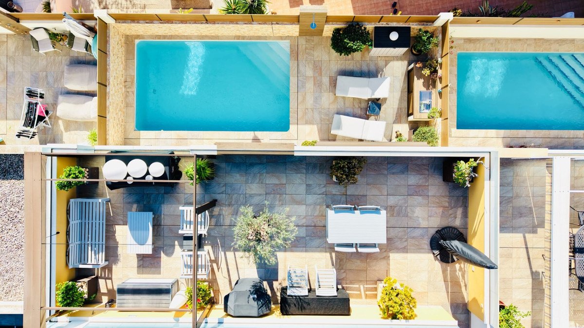 Aerial view of a rooftop terrace with two swimming pools, outdoor seating areas, sun loungers, and dining table.