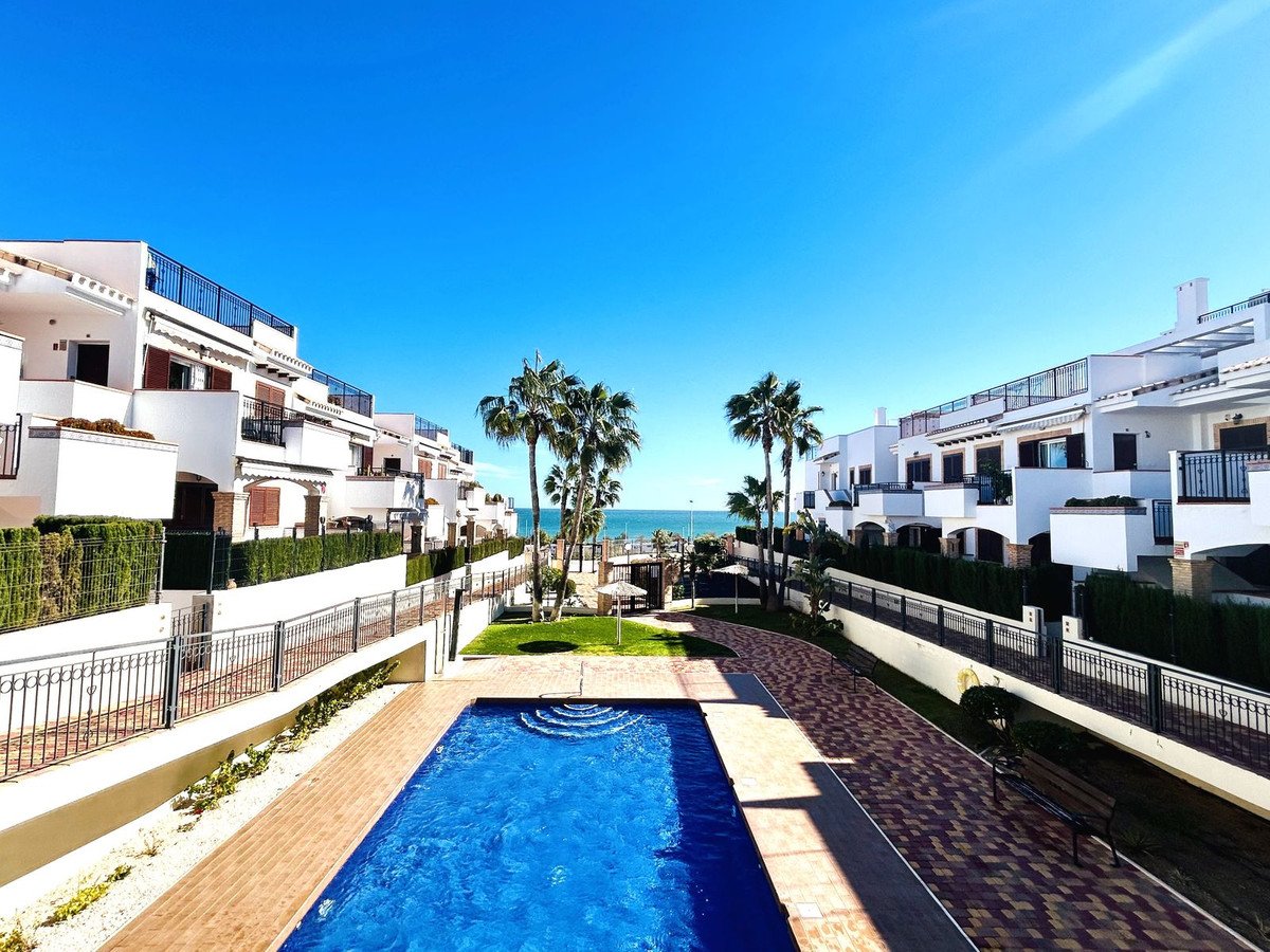 Modern white townhouses with balconies and a swimming pool, palm trees, and the sea in the background under a clear blue sky.
