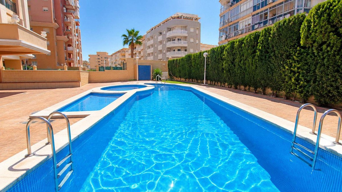 A bright blue swimming pool with a jacuzzi area and metal ladders, surrounded by a paved deck and lush green hedges.
