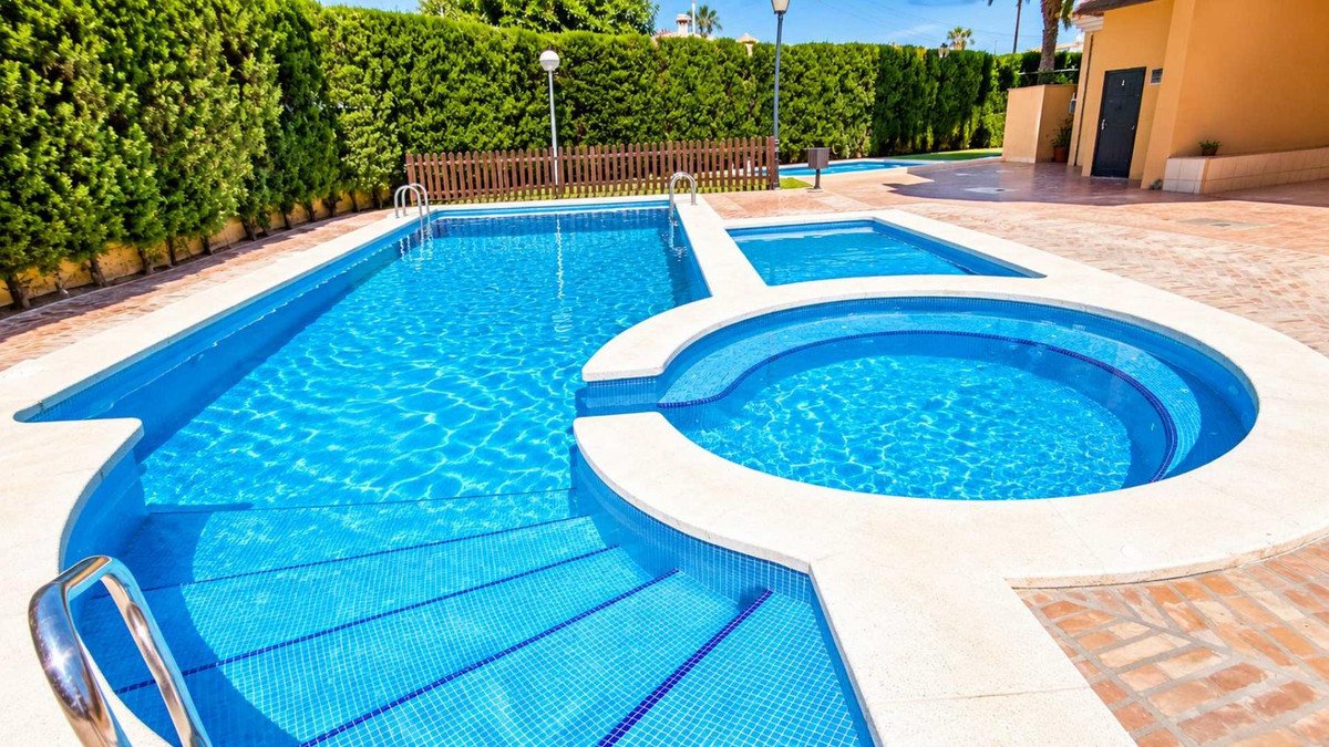 A blue swimming pool with a jacuzzi and steps, surrounded by a paved area and green hedges.