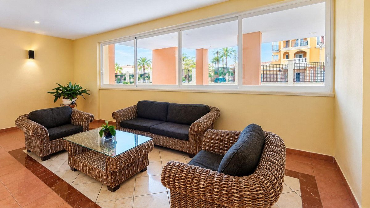 A bright seating area with wicker sofas and chairs, a glass coffee table, and a view of palm trees and buildings through large windows.