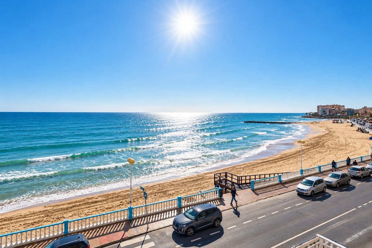 A wide sandy beach with gentle waves under a bright sunny sky, with a street and cars in the foreground and buildings in the distance.