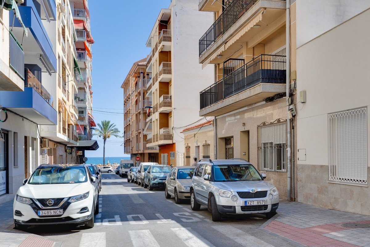 Street view leading to the sea with parked cars and apartment buildings under a clear blue sky.