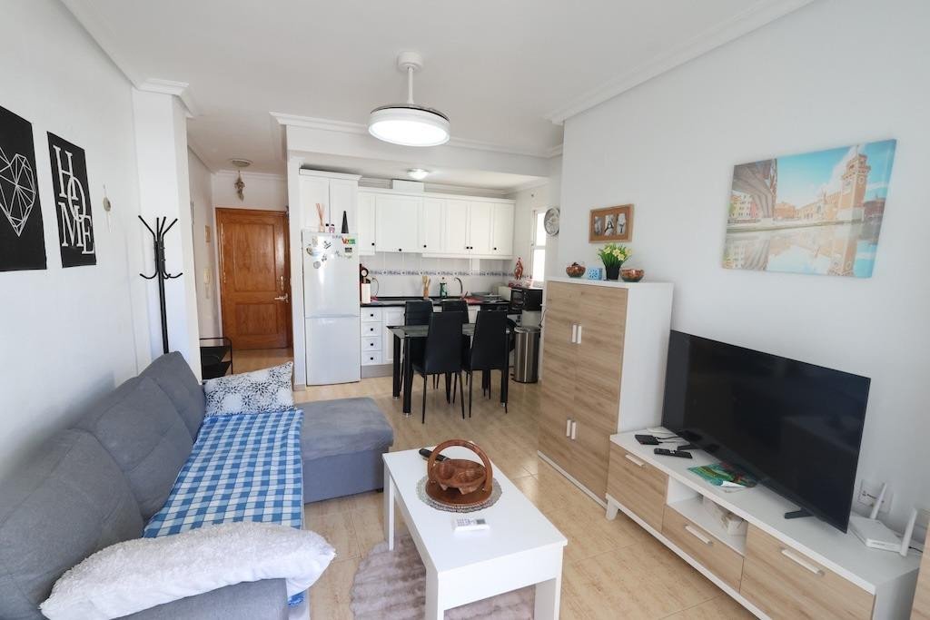 A modern living room with a grey sofa and plaid blanket, adjacent to a dining area and a white kitchen with modern appliances.