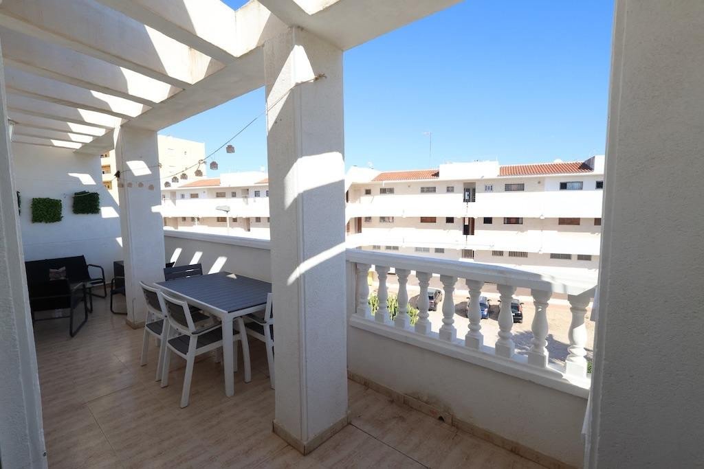 A covered balcony with a dining table, chairs, and a sofa, overlooking buildings under a clear blue sky.