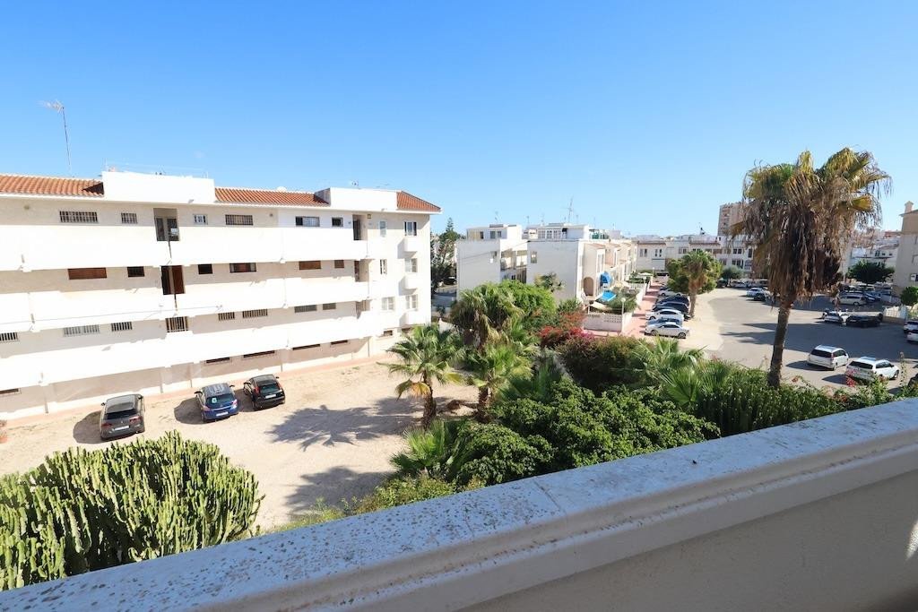 View from a balcony overlooking apartment buildings, parking lot with cars, and palm trees under a clear blue sky.