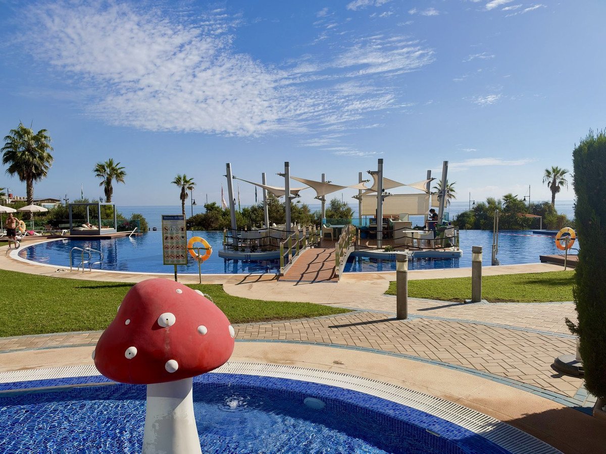 Outdoor swimming pool area with a bar, lounge beds, palm trees, and a distant sea view.