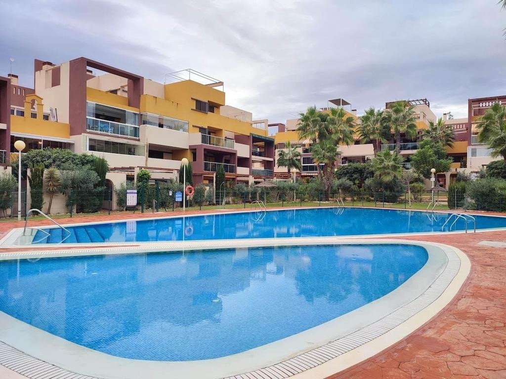 Outdoor swimming pool with modern apartment buildings and palm trees in the background on a cloudy day.