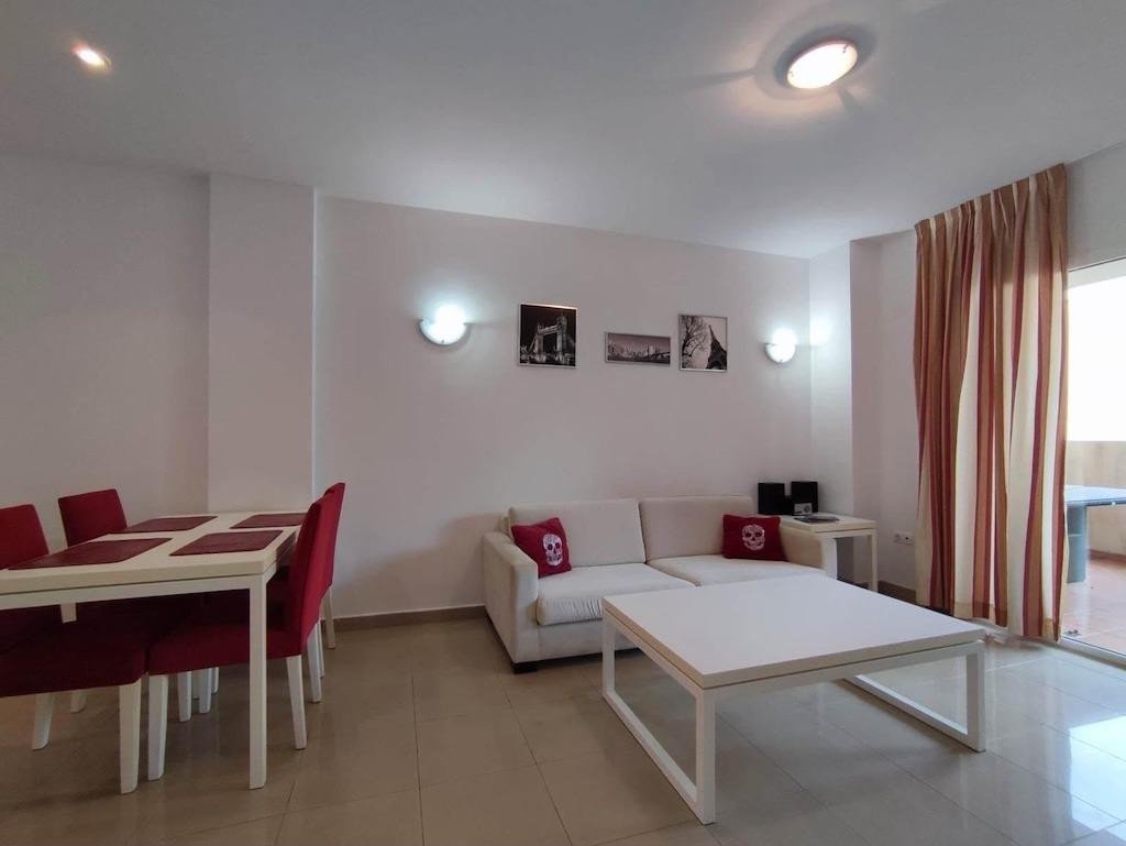 Bright living and dining area with a sofa, coffee table, dining table with red chairs, and balcony access through sliding doors.