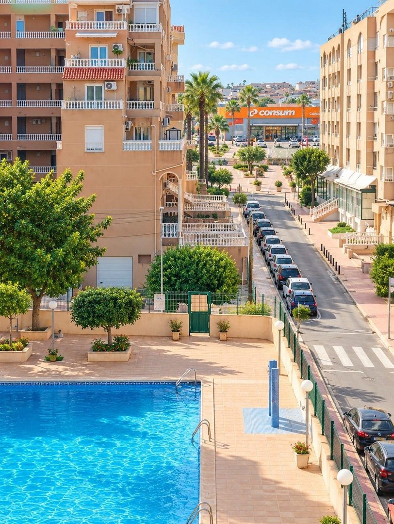 Bright blue swimming pool next to apartment buildings with a street and shops in the background. Sunny day.