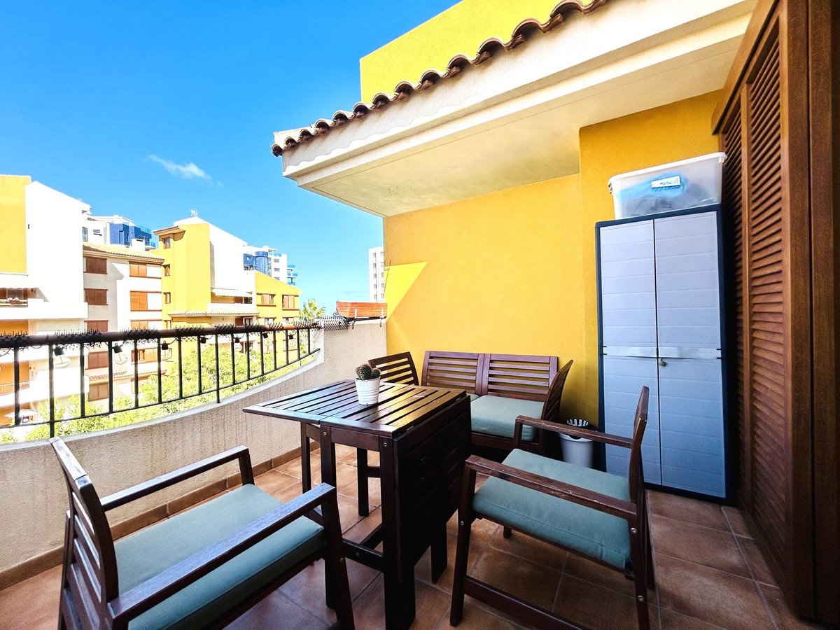 Balcony with wooden table, chairs, and bench, overlooking buildings and trees under a blue sky.