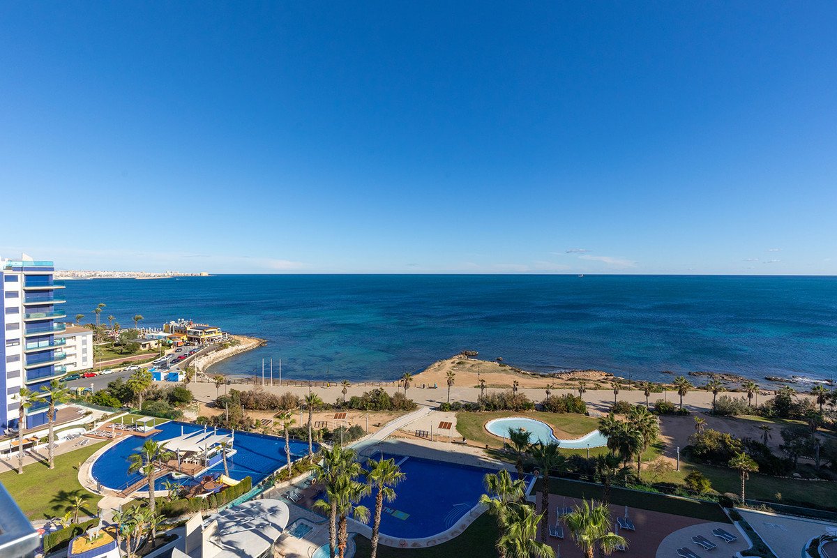 Panoramic view from a balcony of a resort with multiple pools and palm trees overlooking the blue sea and coastline.