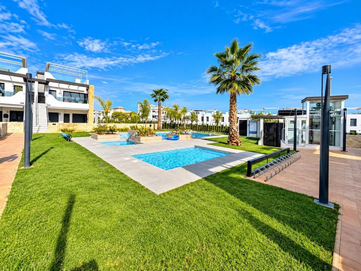Modern apartment complex with multiple swimming pools, well-kept lawns, palm trees, and an outdoor elevator.
