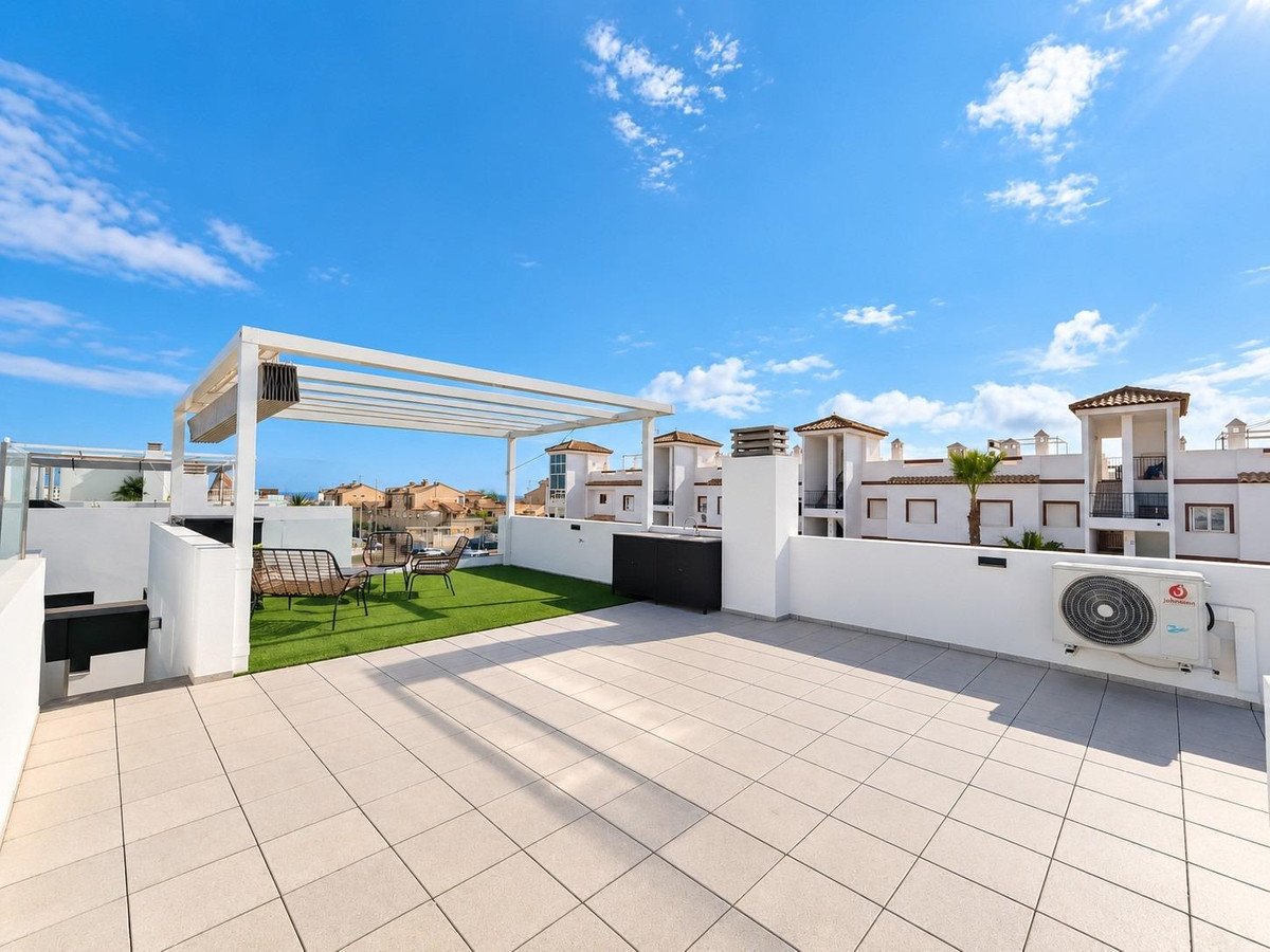 A spacious rooftop terrace with tiled flooring, artificial grass, seating area, pergola, and distant buildings under a blue sky.
