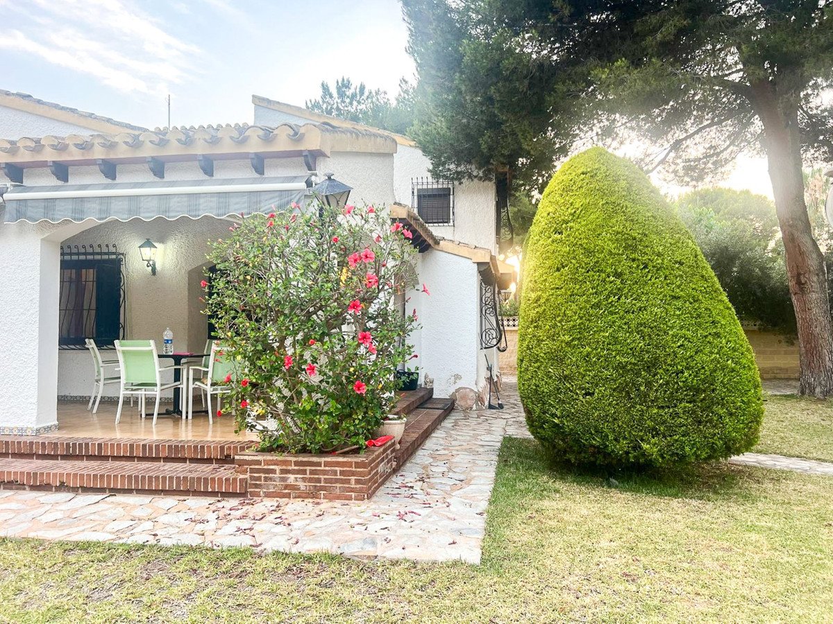 A covered patio with a dining set sits beside a lush green garden with flowering bushes and a large sculpted hedge.