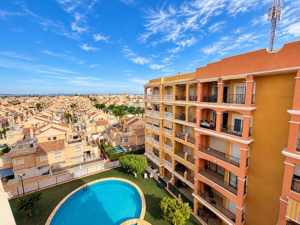 View from apartment balcony showing a resort with a swimming pool and surrounding townhouses under a blue sky.