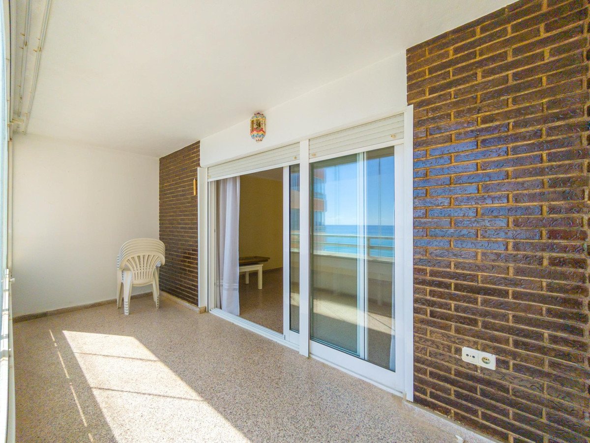 Balcony with sea view, sliding glass doors, white chairs, and a brick accent wall. Sunlight streams in.