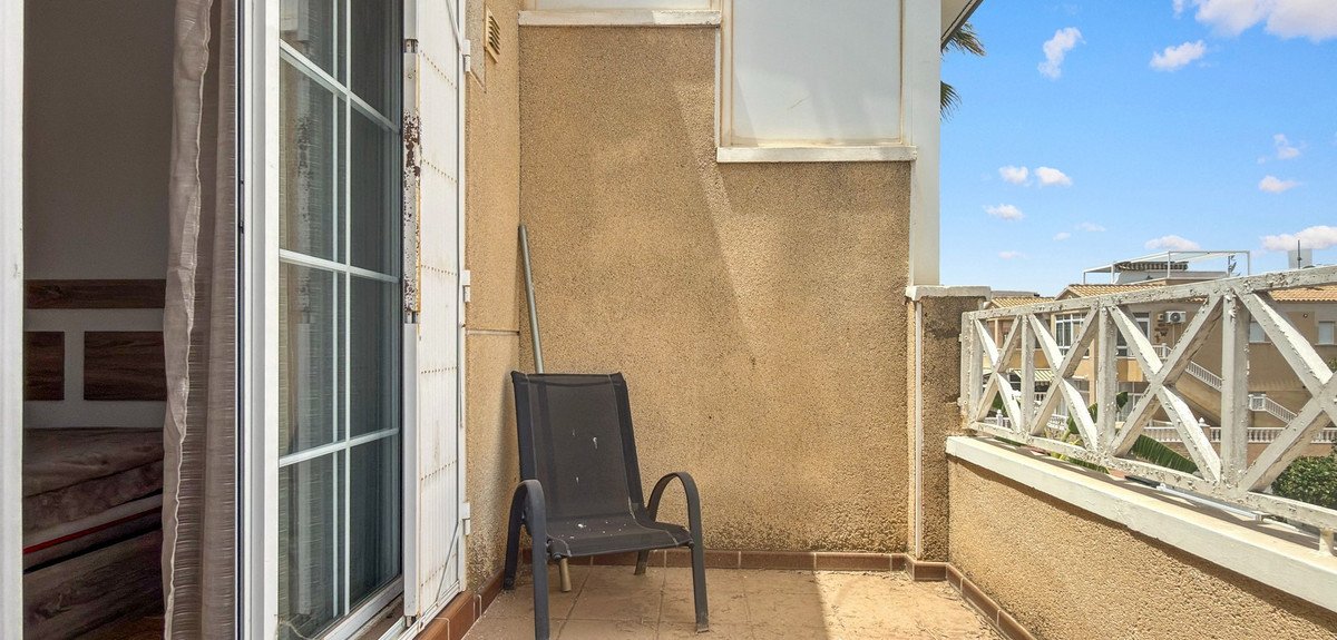 A simple balcony with a black chair and a view of buildings and the sky on a sunny day.