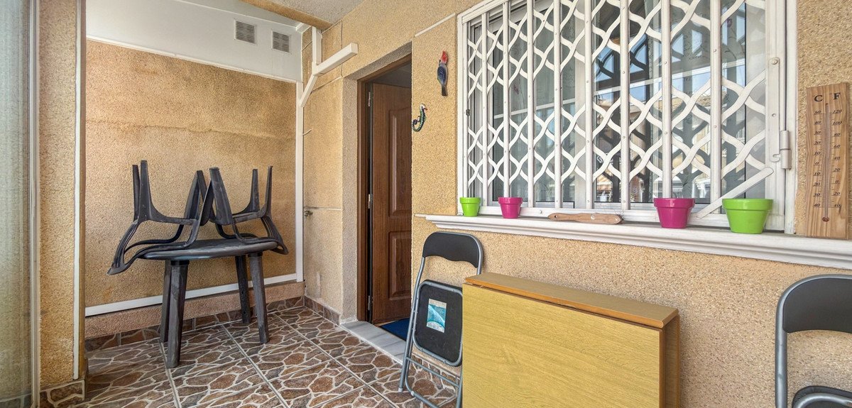 A covered balcony with a table and chairs, a closed window with decorative bars, and colorful flower pots.
