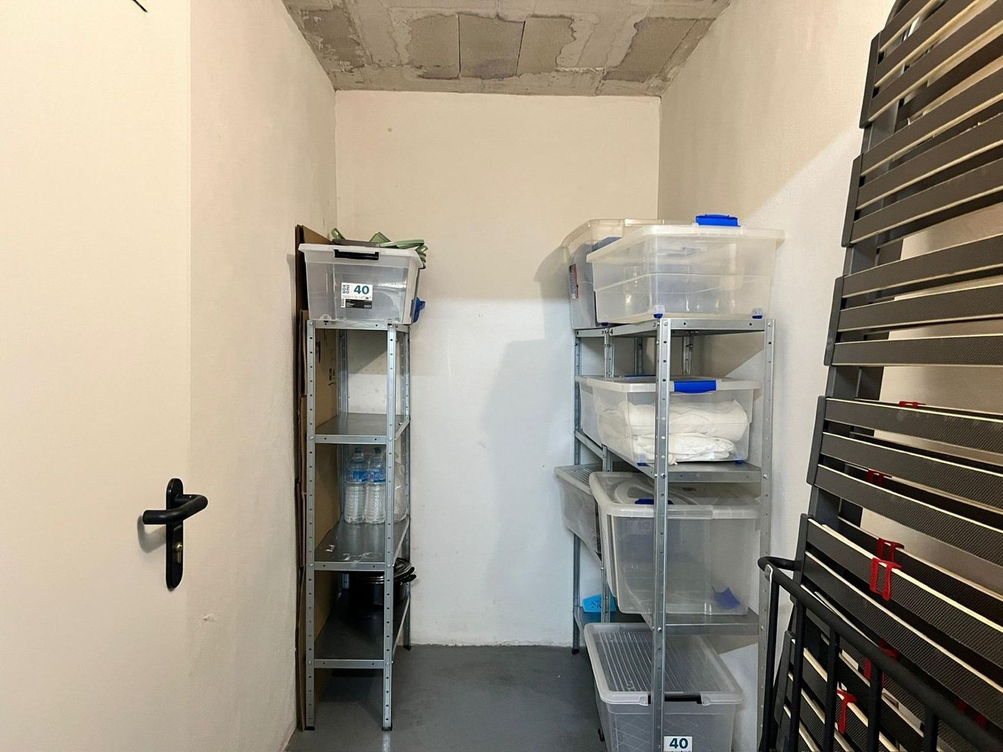 Kleine opslagruimte met metalen rekken, plastic bakken en een deur.