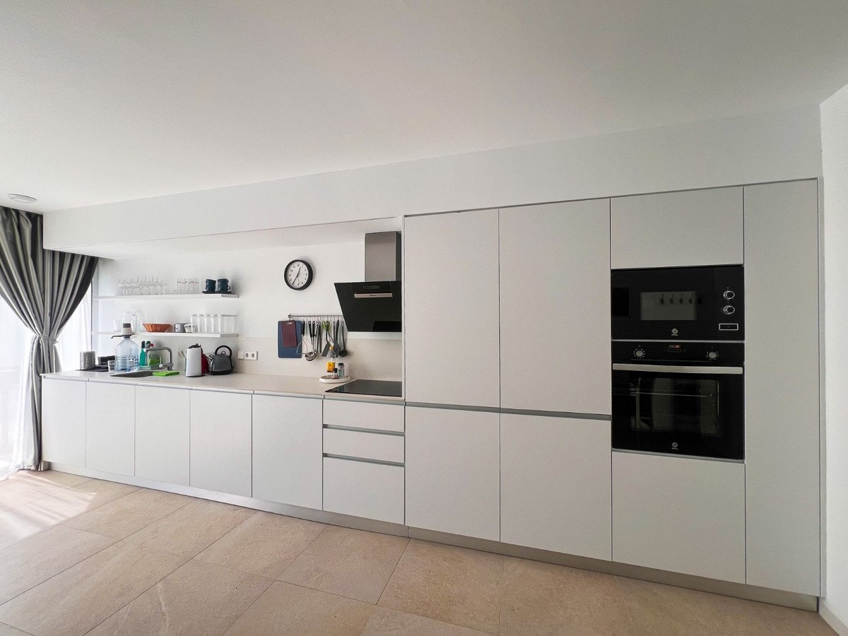 A modern white kitchen with clean lines, integrated appliances including an oven and microwave, and a sink area.
