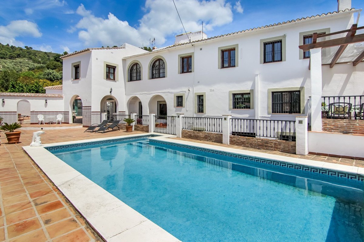 A white villa with a large swimming pool, lounge chairs, and a sunny terrace in a hilly landscape.