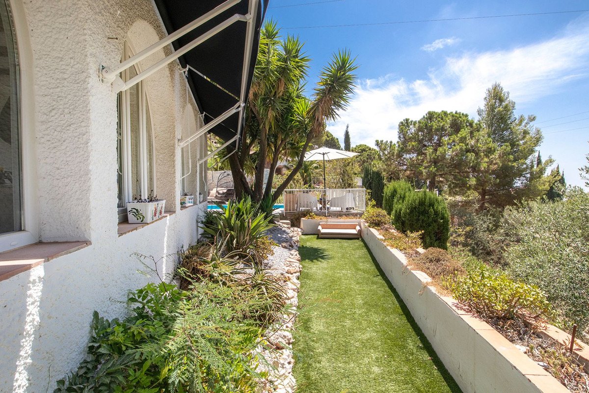 A sunny garden with artificial grass, a pool, and a terrace with outdoor seating, surrounded by lush greenery and trees.