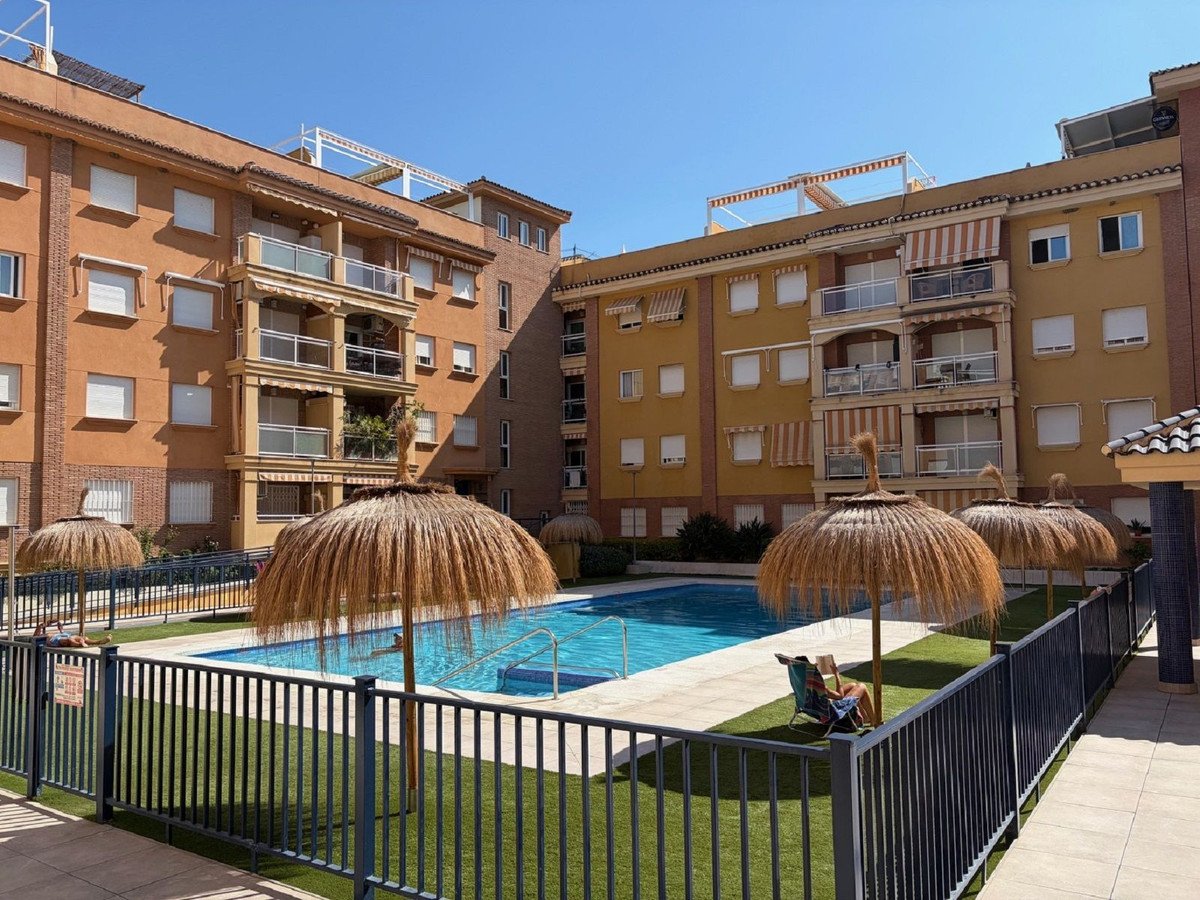 Apartment buildings overlook a swimming pool with thatched umbrellas and loungers on a sunny day.