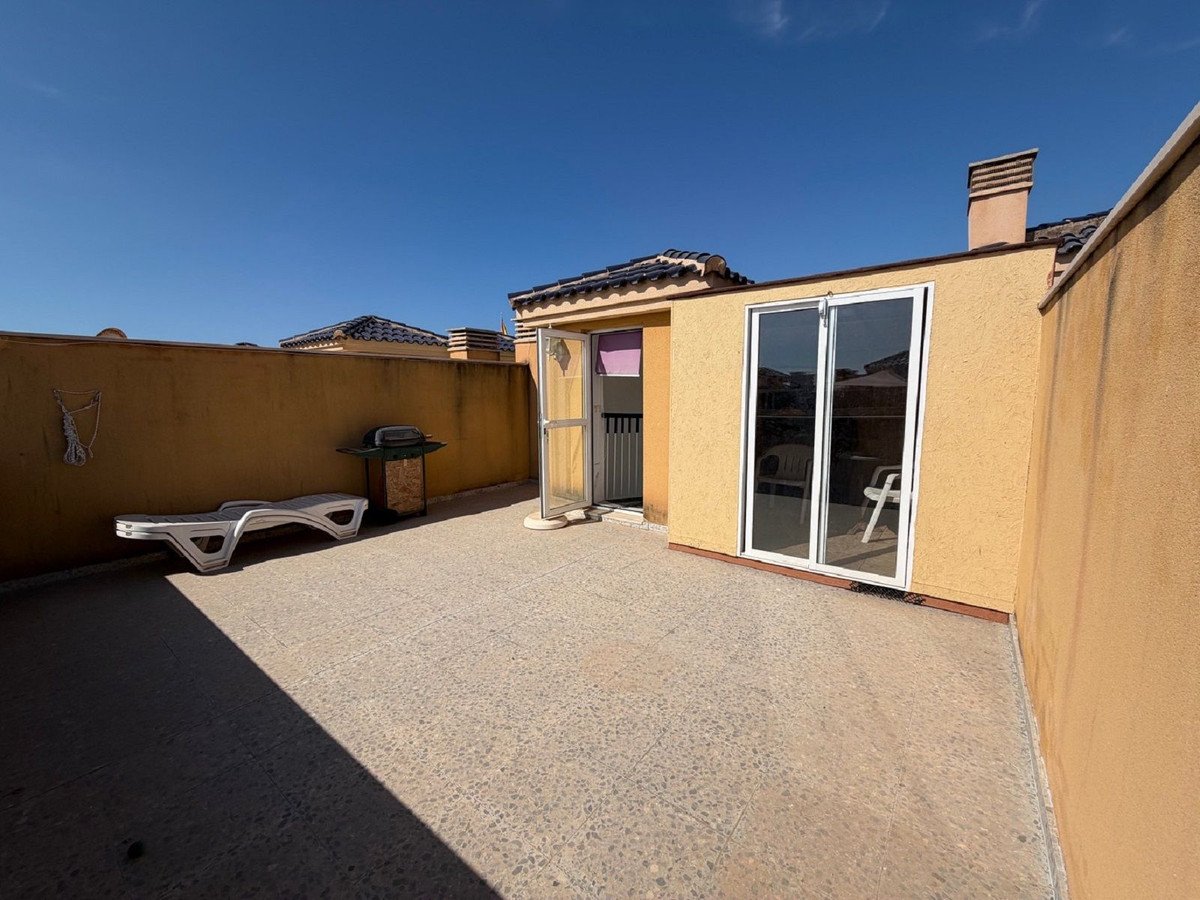A sun-drenched outdoor terrace with a lounger, grill, and sliding glass doors leading indoors. Clear blue sky overhead.