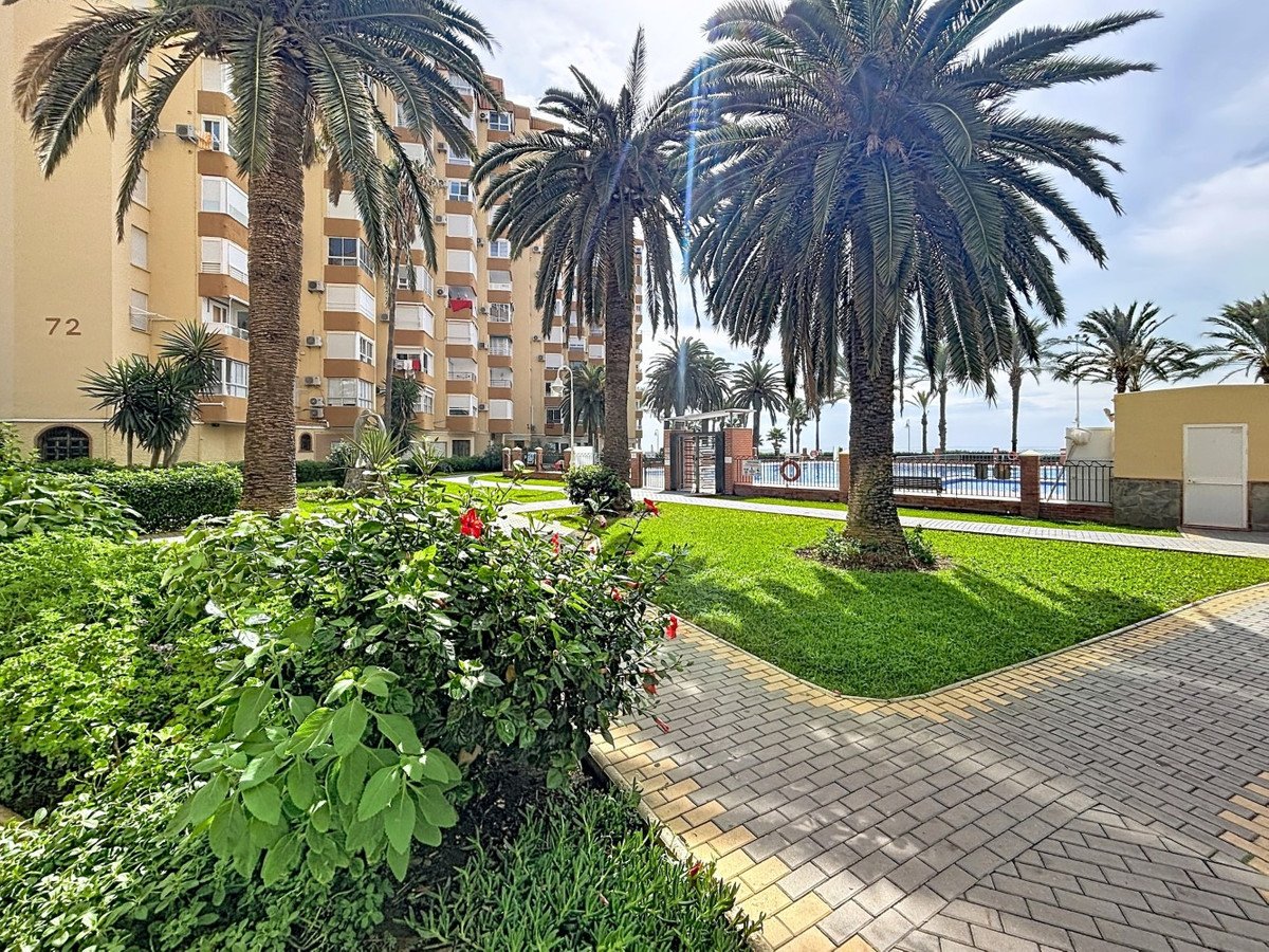 Apartment building exterior with palm trees, a swimming pool, and a view of the sea in the background.