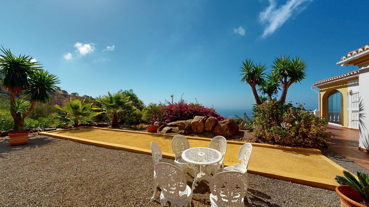 An outdoor seating area with white wrought iron furniture, a bocce ball court, palm trees, and a distant view of the sea.