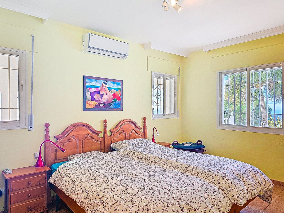 A sunny bedroom with two beds, air conditioning, and windows offering a view of the ocean and palm trees.