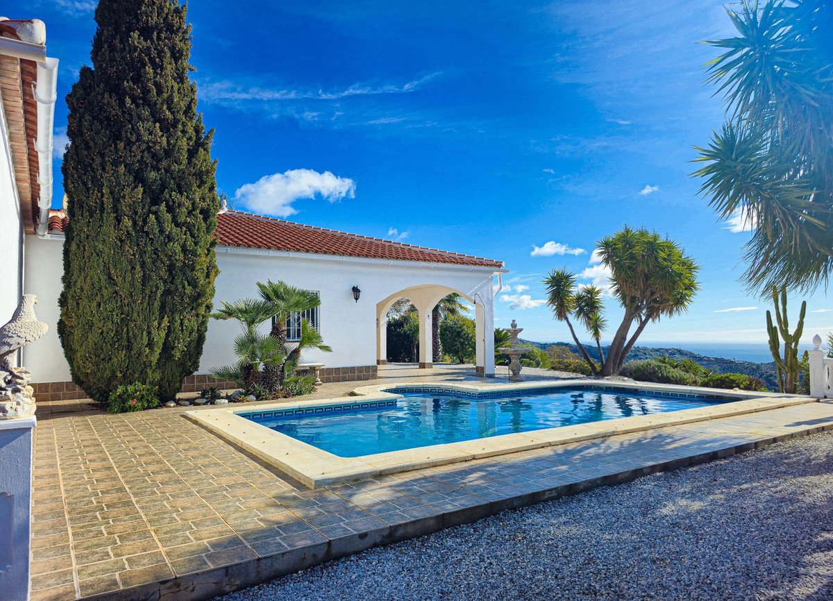 Exterior view of a white villa with a swimming pool, sea view, and lush greenery under a blue sky.