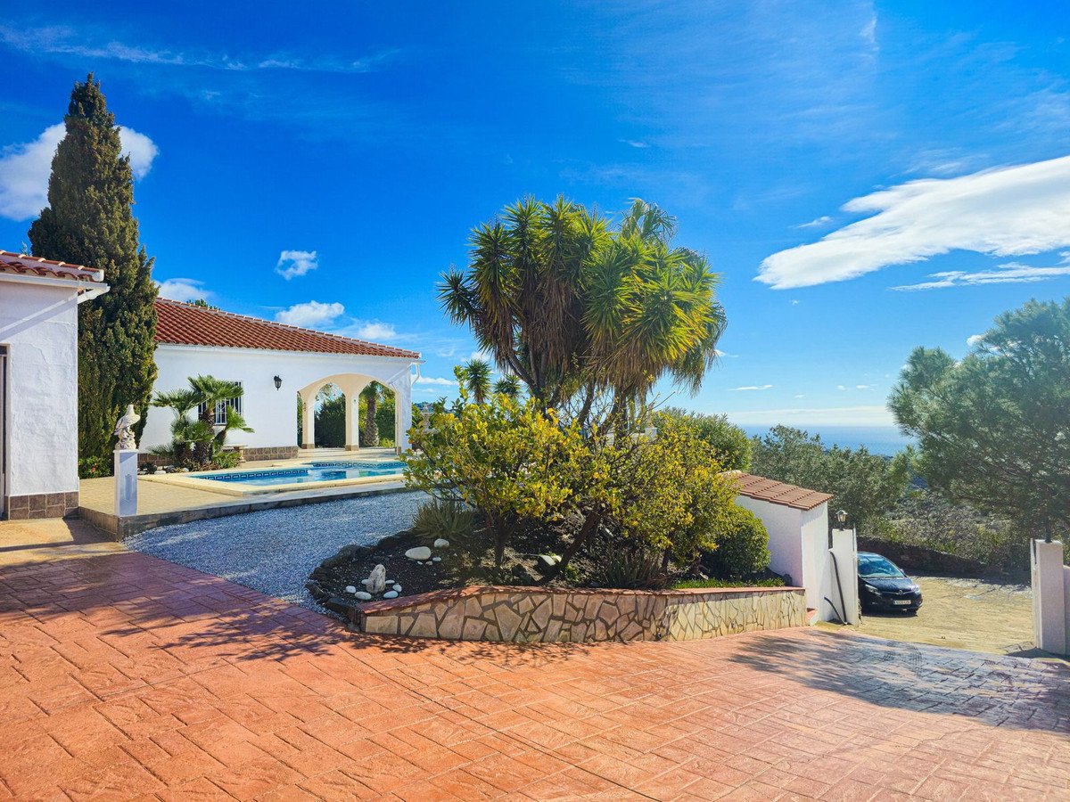 White villa with a blue swimming pool, lush greenery, and a distant view of the sea under a bright blue sky.