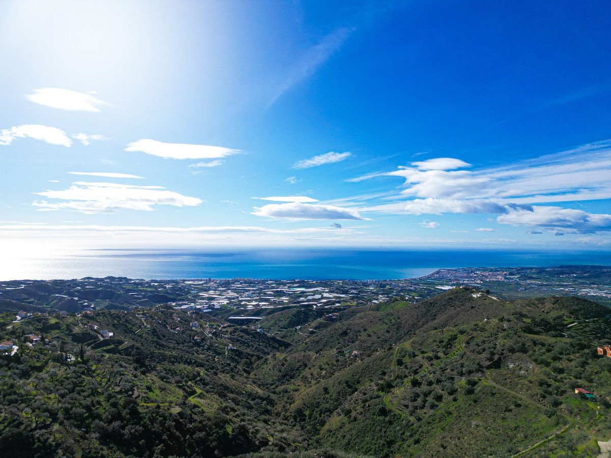 Panoramic view of a hilly landscape with scattered houses, a distant town, and the sea under a bright blue sky.