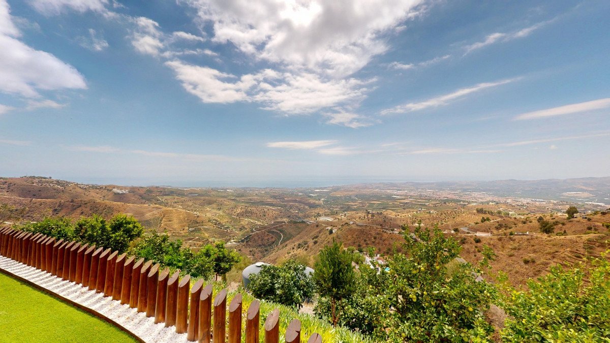 Expansive view of a hilly landscape with distant sea, featuring lush greenery and a modern wooden fence in the foreground.