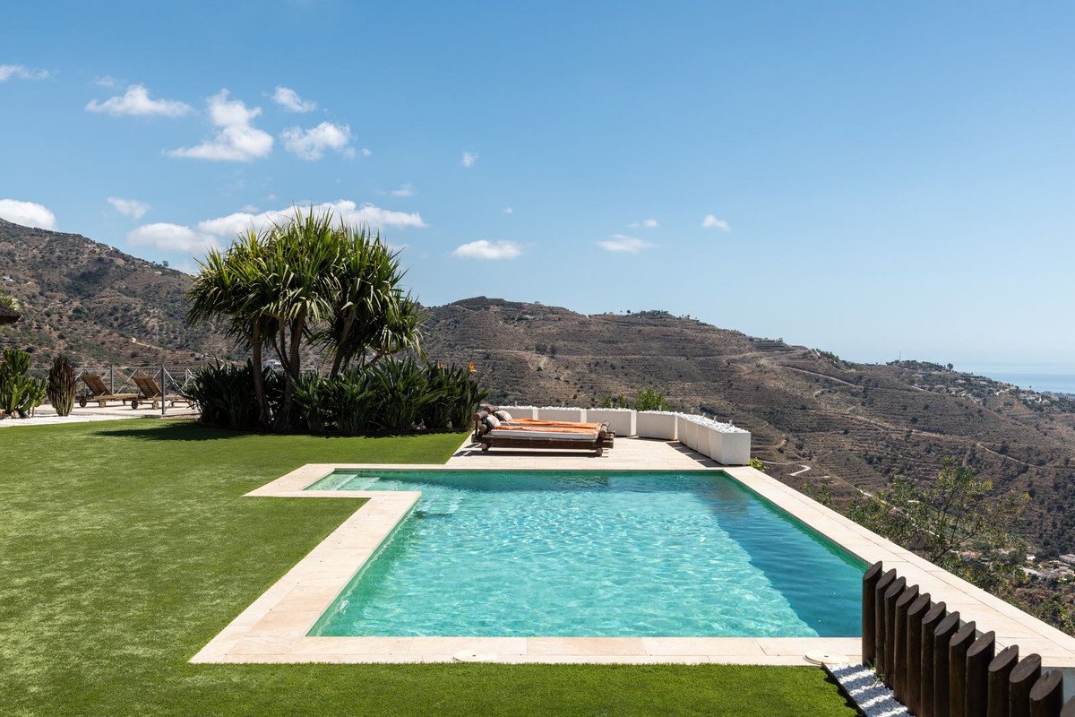 An infinity pool with a daybed and lounge chairs overlooks a stunning mountain landscape and distant sea.