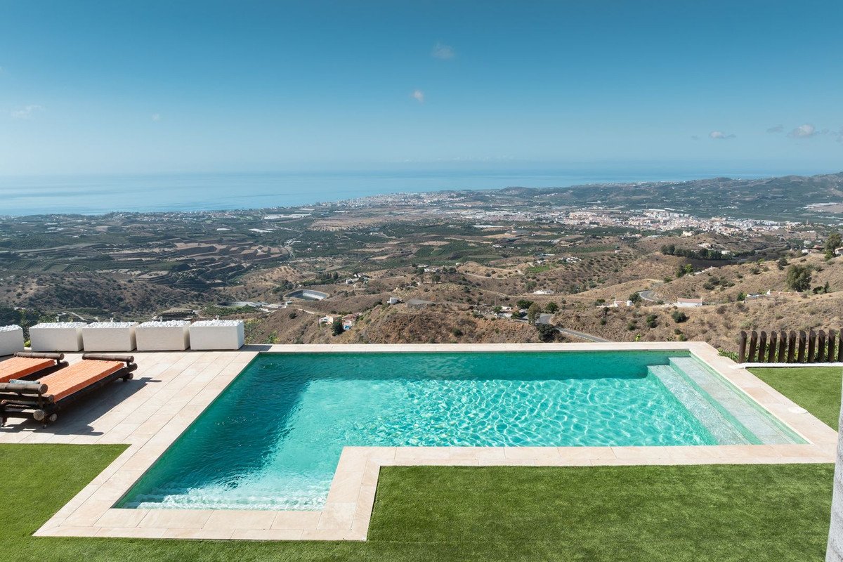 An infinity swimming pool overlooking a coastal town and the sea, with lounge chairs on the terrace.