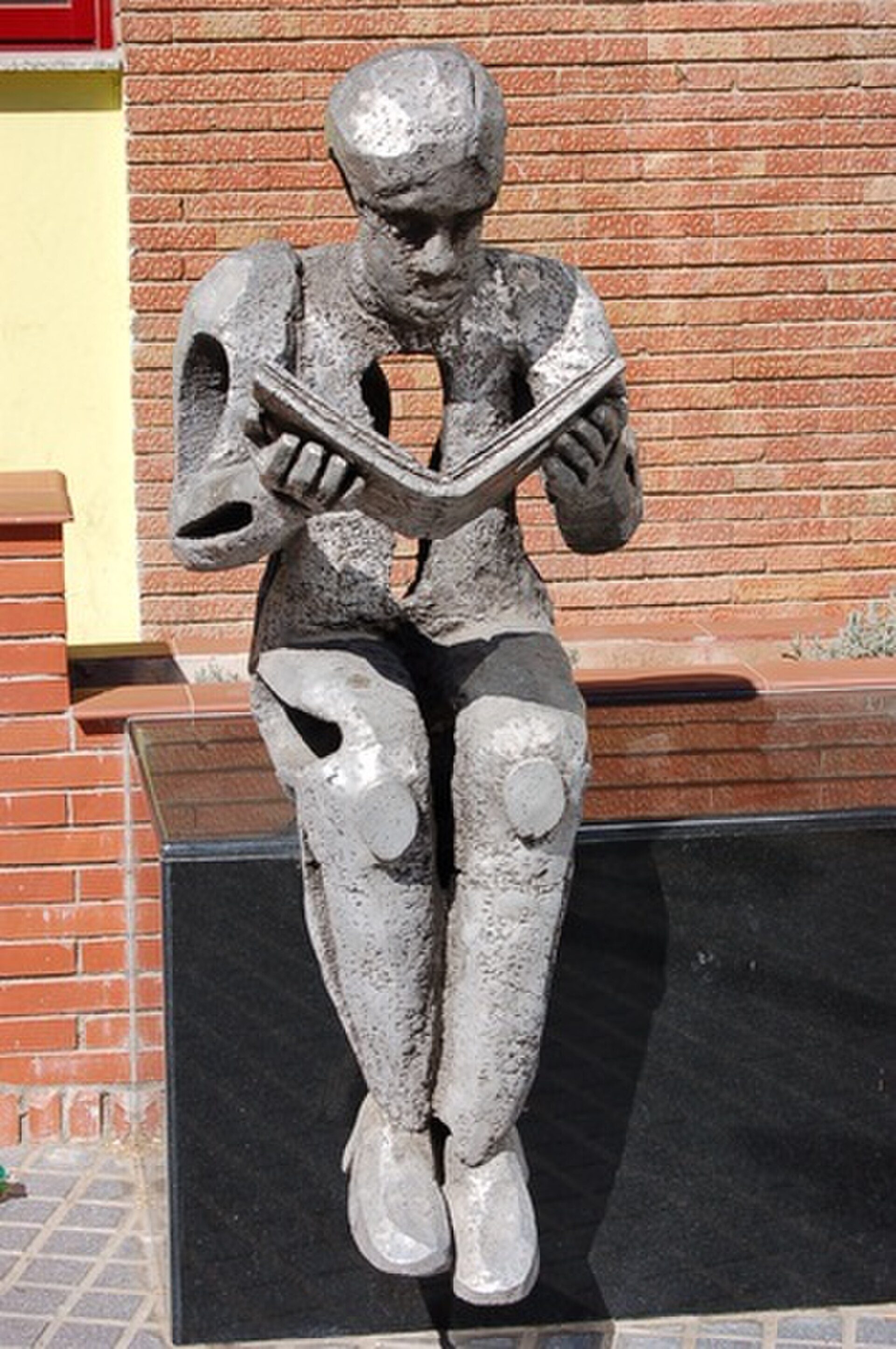 A bronze statue of a person reading a book, sitting on a bench against a brick wall.