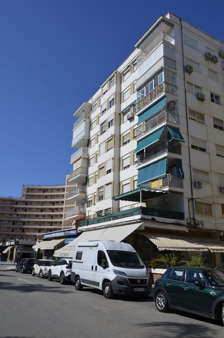 Exterior of a multi-story apartment building with balconies, awnings, and parked cars on the street below.