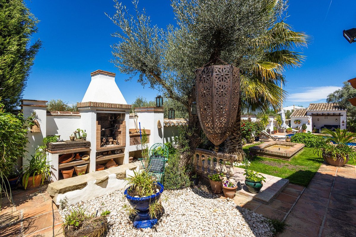 Outdoor kitchen with built-in barbecue, seating area, small fountain, and pool in the background on a sunny day.