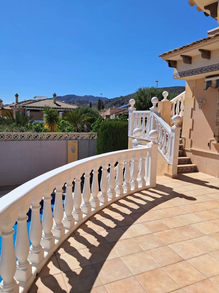 A sunlit patio area with a white balustrade overlooking a blue swimming pool and distant hills.